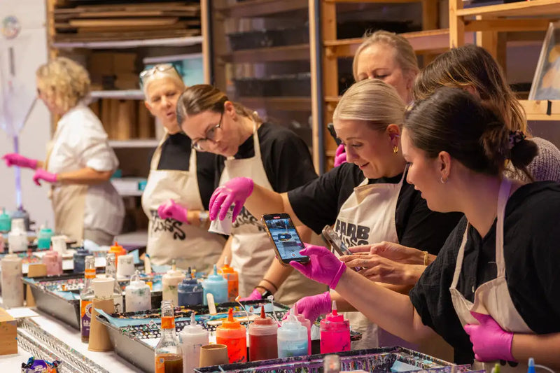 Eine Frau, die rosa Handschuhe und eine weiße Schürze mit dem Aufdruck „garb“ trägt, hält ihr Smartphone hoch, um den Moment festzuhalten, während sie an einem Kunstkurs teilnimmt.