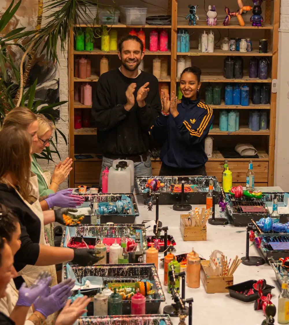 Ein Mann in einem schwarzen Hoodie steht lächelnd mit gefalteten Händen, positioniert hinter einem Tisch, der mit bunten Kunstbedarfsmaterialien und lebendigen Farbbottichen auf Regalen gefüllt ist.